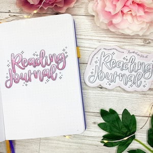 May include: An open notebook with the words "Reading Journal" in pink script, accompanied by a matching sticker. The notebook has dotted pages and a yellow ribbon. Decorative elements include a pink flower and green leaves.