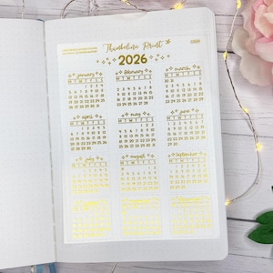 May include: A planner spread featuring a gold foil calendar sticker for 2026. The sticker, labelled "Thumbelina Print", is divided into monthly calendars. The planner pages are white with a dotted grid, and a pink flower is visible.