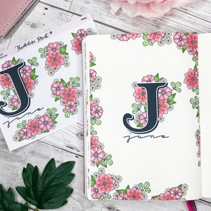 May include: A sheet of printable stickers featuring a floral design with the letter "J" and the word "June" in black script. The stickers are on a white background with a pink and green floral border.