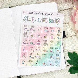 May include: A self-care bingo card with a rainbow gradient background, featuring activities like "Take a nap" and "Enjoy a bath." The card is on a white notebook page, with the words "SELF-CARE BINGO" at the top.