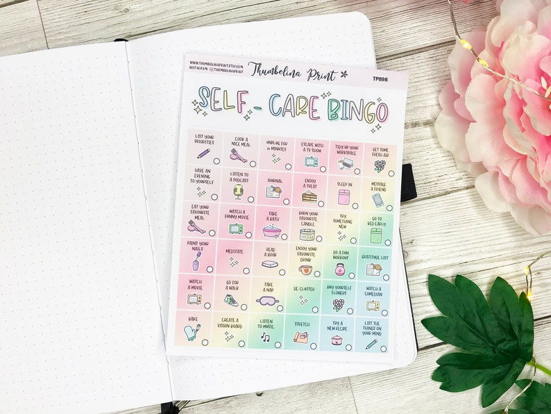 May include: A self-care bingo card with a rainbow gradient background, featuring activities like "Take a nap" and "Enjoy a bath." The card is on a white notebook page, with the words "SELF-CARE BINGO" at the top.