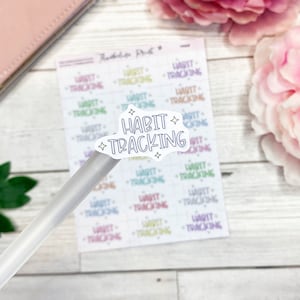 May include: A sheet of colourful "Habit Tracking" stickers with a single sticker on a pen. The sticker is white with black text and stars.
