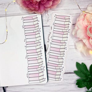 May include: A notebook and a sticker featuring stacks of books with pink and white pages. The notebook is open to a blank page with dot grid paper. The sticker has the text "Threshold Print". Pink flowers and fairy lights are in the background.