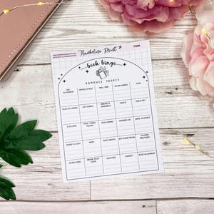 May include: A printable book bingo card with a black and white design. The card features a list of common romance tropes, such as "fake engagement" and "enemies to lovers".