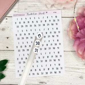 Op de afbeelding: Een wit vel papier met een raster van nummers van 1 tot en met 31. Drie witte ronde stickers met zwarte nummers 19, 20 en 21 zijn geplaatst op een doorzichtige plastic staaf.