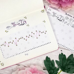 May include: A pink and white sleep log planner with a line graph and stars. The planner has the text "Sleep Log" written in cursive. The planner is on a white wooden surface.