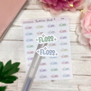 May include: Two colorful cloud-shaped stickers with the word "FLOSS" written on them. The stickers are on a clear pen and are being used to decorate a planner.