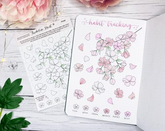 May include: A journal spread with a pink floral design and the words "habit tracking" in pink script. A sheet of stickers with matching floral outlines is next to the journal. The stickers and journal feature cherry blossom designs.