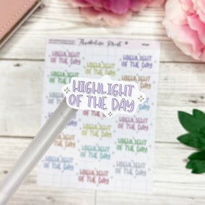 May include: A sheet of stickers with the text "HIGHLIGHT OF THE DAY" in a cloud-shaped design. The stickers are in various colors, including purple, green, and yellow. A clear pen is positioned over the stickers.
