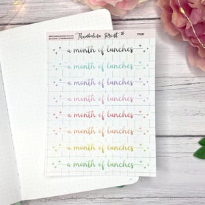 May include: A sticker sheet with the text "a month of lunches" in various colors, including black, teal, purple, pink, orange, and yellow. The stickers are on a grid background and are placed on an open notebook.