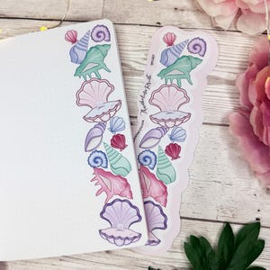 May include: A white dot grid notebook open to a page featuring a vertical border of colorful seashell illustrations in shades of pink, purple, and teal. Beside the notebook is a matching die-cut sticker sheet with the text Thumbelina Print.