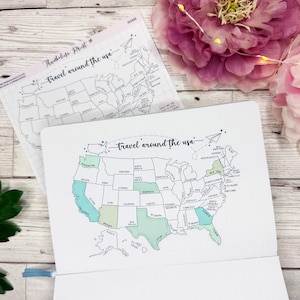 May include: Two white notebooks with a map of the USA, titled "Travel around the usa." The map is outlined in black with some states colored in shades of green and blue. The notebooks are on a light wood surface with flowers.