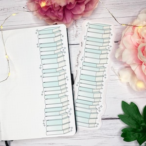 May include: A notebook and a bookmark with a design of stacked books in light blue and white. The bookmark has the text "Read.Love.Repeat." and star decorations. Pink flowers and fairy lights are in the background, creating a decorative scene.