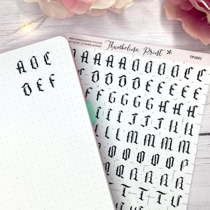 Könnte beinhalten: Ein weißes, gepunktetes Notizbuch mit den Buchstaben A, B, C, D, E und F in schwarzer gotischer Schrift. Daneben befindet sich ein Bogen mit gotischen Alphabetaufklebern mit dem Text "Thumbelina Print" und "TP1461".