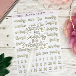 Könnte beinhalten: Ein Blatt weißes Aufkleberpapier mit goldenen und weißen Buchstaben. Die Aufkleber zeigen die Wochentage, die Zahlen von 1 bis 31 und die Worte "Thumbelina Print".