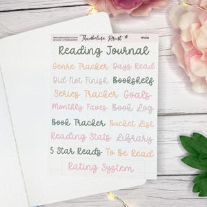May include: A sticker sheet with the words "Reading Journal" and other phrases like "Genre Tracker" and "5 Star Reads" in various colors. The sticker sheet is on a white dotted notebook page, with pink flowers and green leaves in the background.