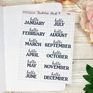 May include: A sheet of stickers featuring the months of the year, each beginning with "hello" in a handwritten style. The stickers are arranged on a grid, with dark blue text. The sheet rests on a dotted notebook page.