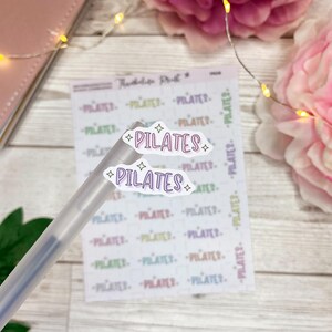May include: Two white stickers with the word "PILATES" written in purple with a pink outline and yellow stars. The stickers are on a clear plastic pen and a sheet of paper with the word "PILATES" printed in various colors.