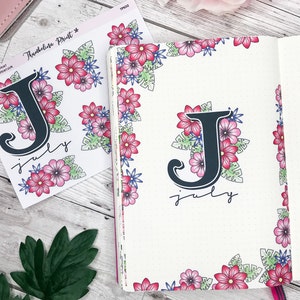 May include: A white planner with a floral border and a black letter J with the word "July" written below it. The planner is open to a blank page. There is a sheet of stickers with floral designs and a black letter J with the word "July" written below it.