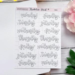 May include: A sheet of stickers with the days of the week in a handwritten font. Each day is written in a cursive style with small star accents. The stickers are on a white background with a grid pattern.