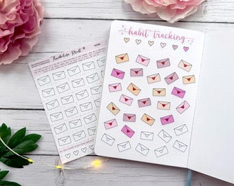 May include: A notebook with the words "habit tracking" and envelope stickers with hearts. A sheet of envelope stickers is next to the notebook. Pink flowers are in the background.