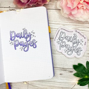 May include: A white notebook with a purple and white "Daily Pages" title written on the left page. A sticker with the same text in black is on the right side of the notebook.