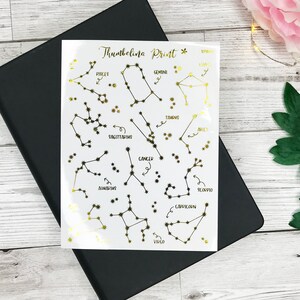May include: A sheet of gold foil constellation stickers with the names of each constellation written in black. The constellations include Pisces, Gemini, Taurus, Sagittarius, Cancer, Aquarius, Scorpio, Capricorn, and Virgo.