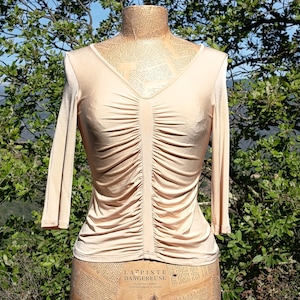 Peut inclure: Haut beige à manches longues avec un col en V et des détails froncés au centre. Le haut est présenté sur un mannequin avec une impression de journal vintage. L'arrière-plan présente un feuillage vert et un ciel bleu.