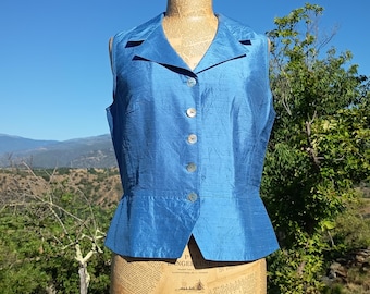 May include: A sleeveless, blue vest with a textured appearance. The vest features a collared neckline, a button-down front, and a slightly flared peplum. The garment is displayed on a mannequin against a backdrop of a blue sky and green foliage.