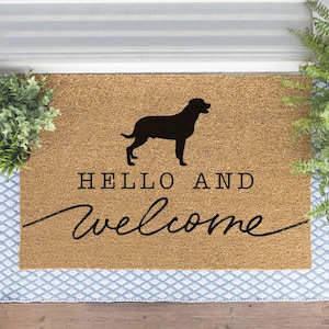 Op de afbeelding: Een bruine kokosmat met de tekst "Hello and welcome" in zwarte cursieve letters. Boven de tekst staat een zwart silhouet van een hond.