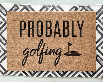 May include: A brown coir doormat with the text "PROBABLY golfing" in black lettering and a black silhouette of a golf hole with a flag.