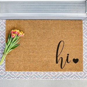 Puede incluir: Una alfombra de coco marrón con la palabra "hi" en letras cursivas negras y un pequeño corazón negro.