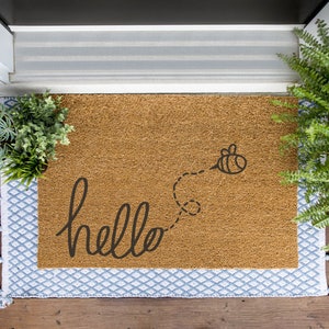 Puede incluir: Una alfombra de coco marrón con la palabra "hello" escrita en letra cursiva y una abeja en blanco y negro volando sobre la palabra. La alfombra está rodeada por un borde estampado azul y blanco con borlas.
