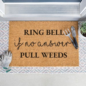 May include: A brown coir doormat with the text "RING BELL if no answer PULL WEEDS" in black lettering. Two gardening tools are placed on the mat.
