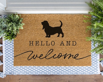 May include: A brown coir doormat with the text "HELLO AND welcome" in black script font. A black silhouette of a basset hound dog is centered above the text.