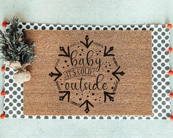 May include: A brown coir doormat with a white and black polka dot border. The doormat has a black graphic that says "baby it's cold outside" in a stylized font.