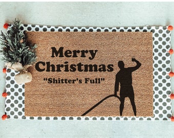May include: A brown coir doormat with the text "Merry Christmas" and "Shitter's Full" in black lettering. A black silhouette of a person holding a drink is on the right side of the mat.