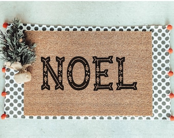 May include: A rectangular doormat with the word "NOEL" in black, decorative lettering. The mat has a brown coir center, a border of white fabric with black polka dots, and red pom-poms. A small Christmas tree is on the left.