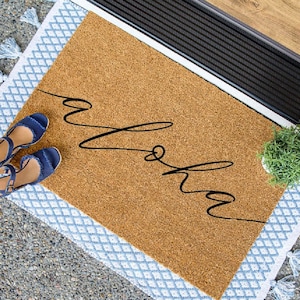 May include: A brown coir doormat with a white and blue patterned border and tassels. The doormat has the word "Aloha" written in black cursive script.