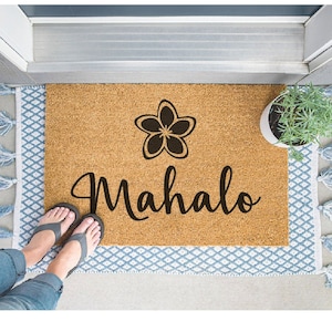 May include: A brown coir doormat with the word "Mahalo" in black cursive lettering and a black silhouette of a plumeria flower.