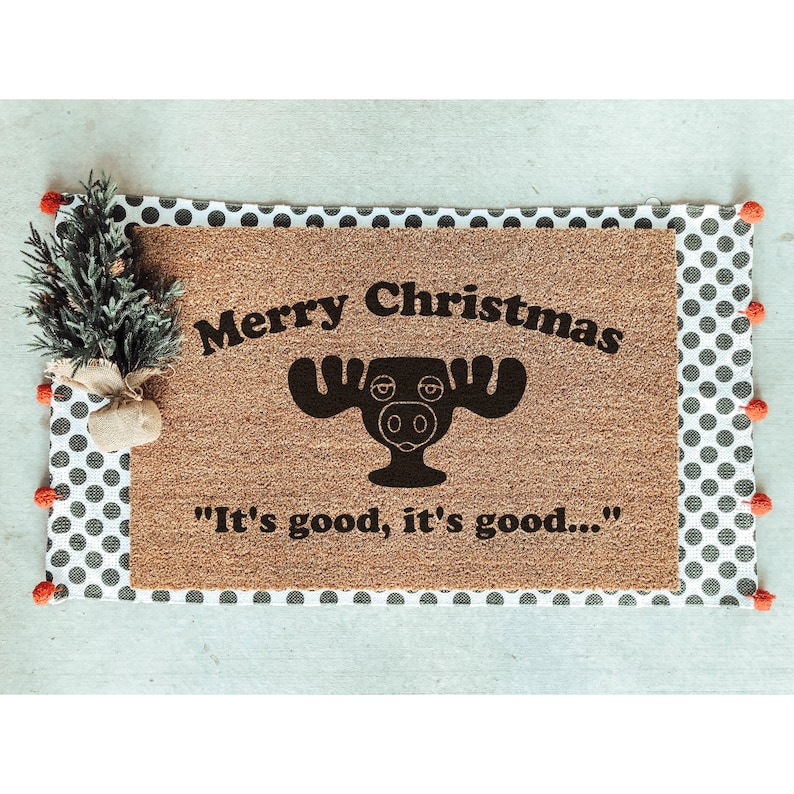 May include: A brown coir doormat with the text "Merry Christmas" and a black moose illustration. The text "It's good, it's good..." is printed below the moose.