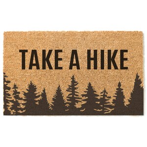 K&ouml;nnte beinhalten: Eine braune Kokosmatte mit dem Text "TAKE A HIKE" in schwarzen Buchstaben. Der untere Teil der Matte zeigt eine Silhouette eines Waldes mit Kiefern.