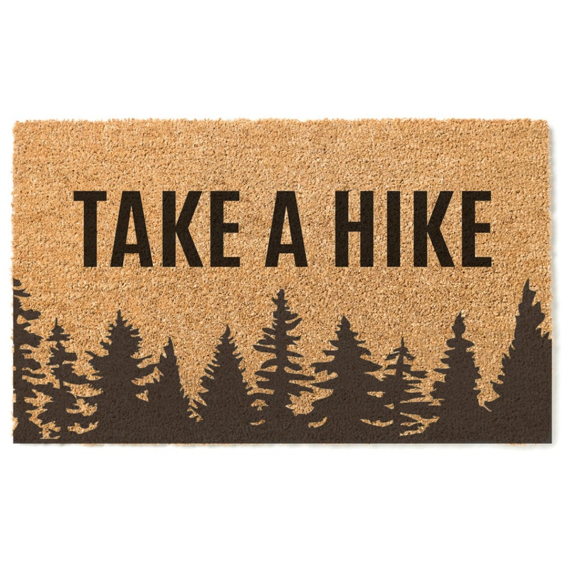 K&ouml;nnte beinhalten: Eine braune Kokosmatte mit dem Text "TAKE A HIKE" in schwarzen Buchstaben. Der untere Teil der Matte zeigt eine Silhouette eines Waldes mit Kiefern.