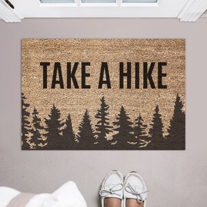 K&ouml;nnte beinhalten: Eine braune Kokosfaser-Fu&szlig;matte mit dem Text "TAKE A HIKE" in schwarzen Buchstaben. Die Fu&szlig;matte hat eine Silhouette eines Waldes mit schwarzen Kiefern.