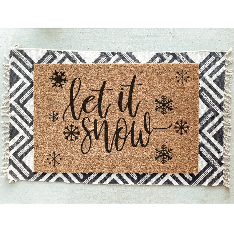 Peut inclure: Un paillasson en fibre de coco brun avec le texte "Let it snow" en lettres cursives noires. Le paillasson est d&eacute;cor&eacute; de plusieurs flocons de neige noirs.