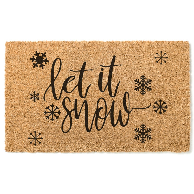Peut inclure: Un paillasson en fibre de coco brun avec le texte "Let it snow" en lettres cursives noires. Le paillasson est d&eacute;cor&eacute; de plusieurs flocons de neige noirs.