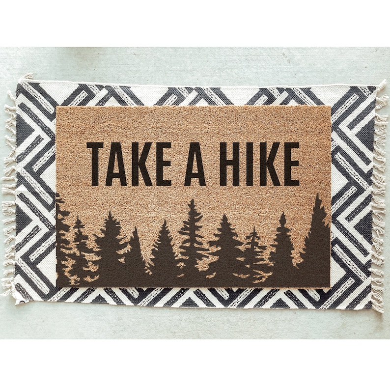K&ouml;nnte beinhalten: Eine braune Kokosfaser-Fu&szlig;matte mit dem Text "TAKE A HIKE" in schwarzen Buchstaben. Die Fu&szlig;matte zeigt eine Silhouette eines Waldes mit Kiefern.