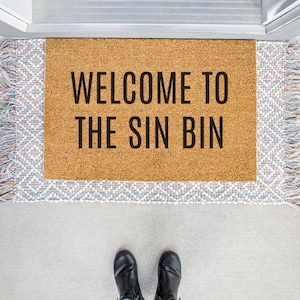 May include: A brown doormat with the text "Welcome to the Sin Bin" in black lettering.