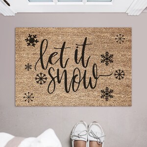 Peut inclure: Un paillasson en fibre de coco brun avec le texte "Let it snow" en lettres cursives noires. Le paillasson est d&eacute;cor&eacute; de petits flocons de neige noirs.
