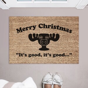 May include: A brown doormat with the text "Merry Christmas" and a black cartoon moose with the text "It's good, it's good..." underneath.